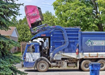An image of a garbage truck picking up a massive waste bin over the top to dump in the waste