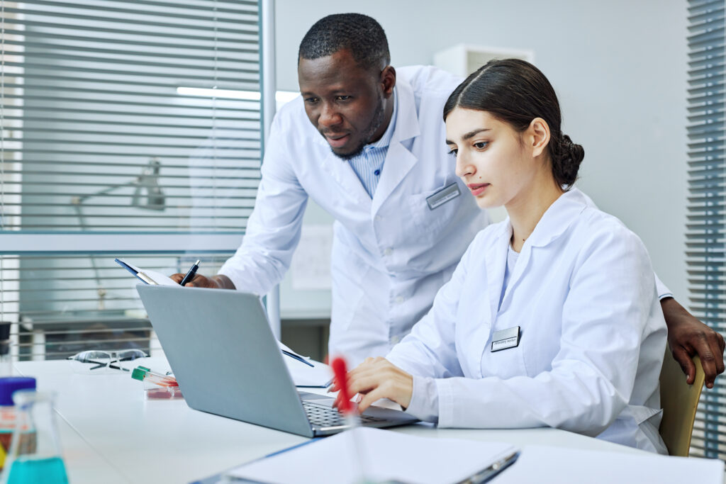 Two researchers, a women seated, typing on a laptop computer and a man looking over her shoulder at the screen. They are both dressed in white lab coats and what appears to be a desk in white laboratory environment.