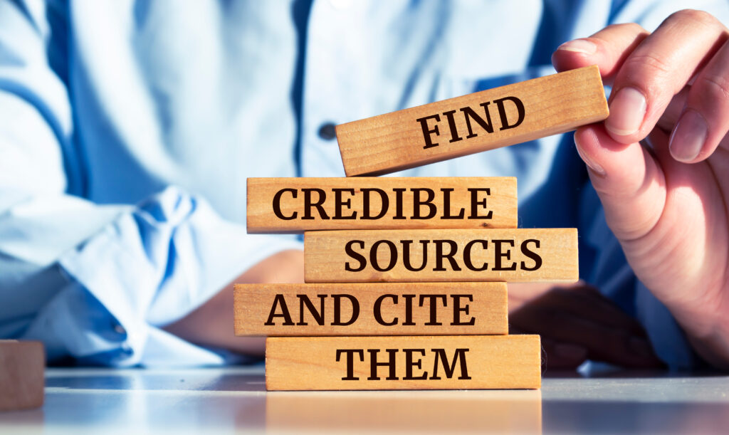 A person putting together a stack of wood blocks that read "Find", "Credible", "Sources" "and Cite" and "Them" atop each other.