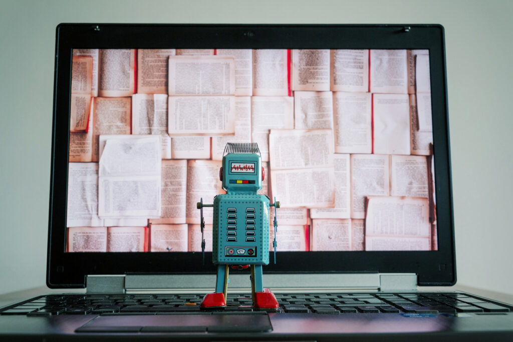 toy robot looking at a laptop screen showing an array of open books