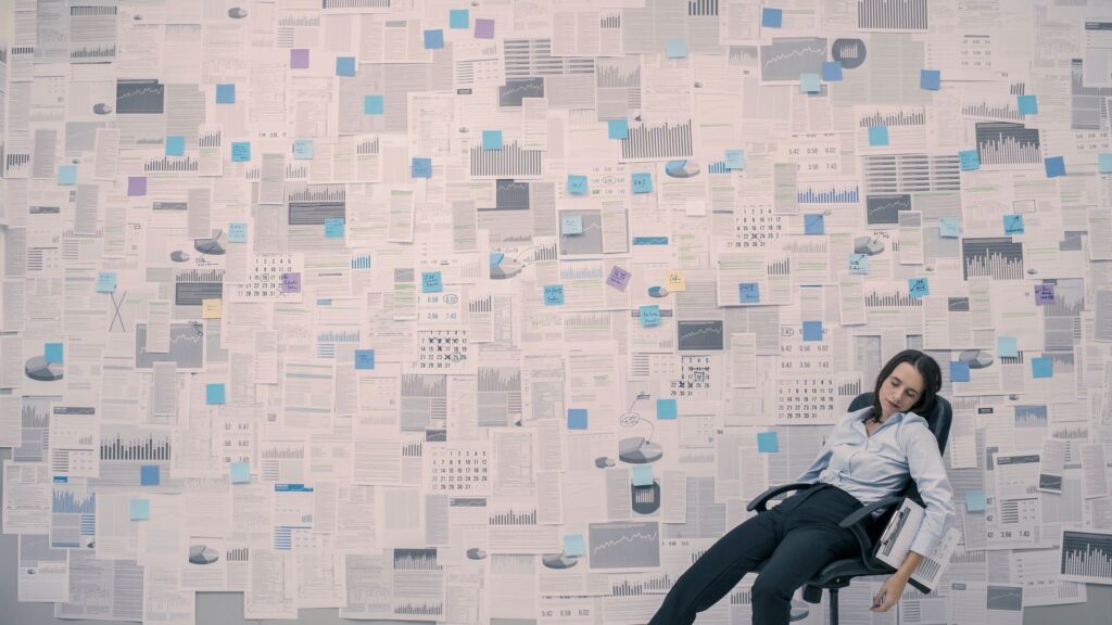 Photo of a wall filled with spreadsheets and statistical data reports. In the lower right corner, a woman sits slumped, apparently exhausted or exasperated, in an office chair with a clipboard under one arm.