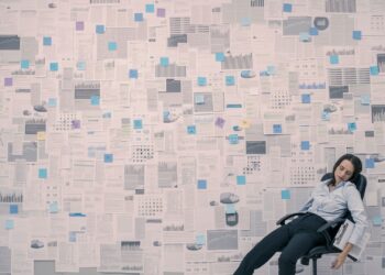 Photo of a wall filled with spreadsheets and statistical data reports. In the lower right corner, a woman sits slumped, apparently exhausted or exasperated, in an office chair with a clipboard under one arm.