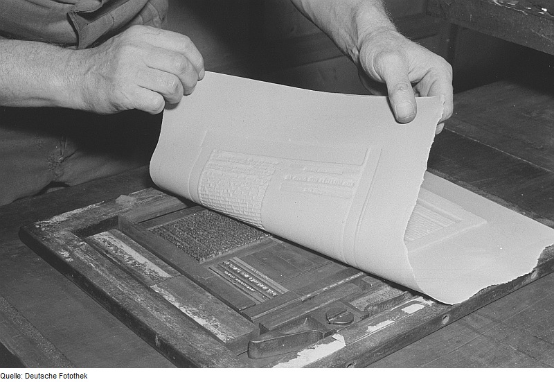 A picture of a flong being created in the 1950s. Paper maché has been pressed onto a metal plate and is being peeled back to show the indentation of the text and images.