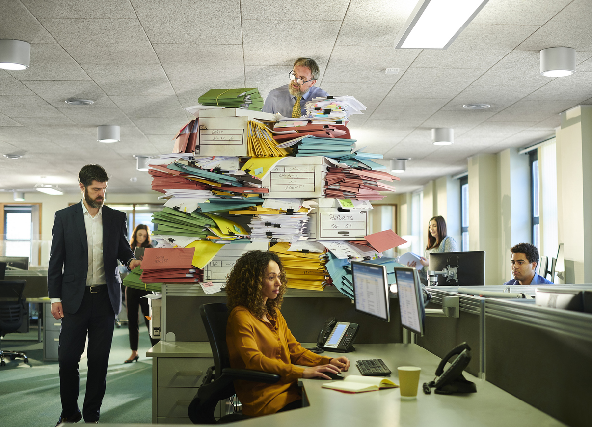 Photo of an older man in a professional office setting, sitting atop a large pile of paperwork. The pile is so tall, the man's head is touching the ceiling. The photo is mean to suggest someone at the later stages of their career rethinking their role in the office.