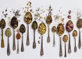 Arrangement of tea and dessert spoons, each spoon with its own blend of tea leaves and other additions, spilling out onto a white background demonstrating variety in tea types.