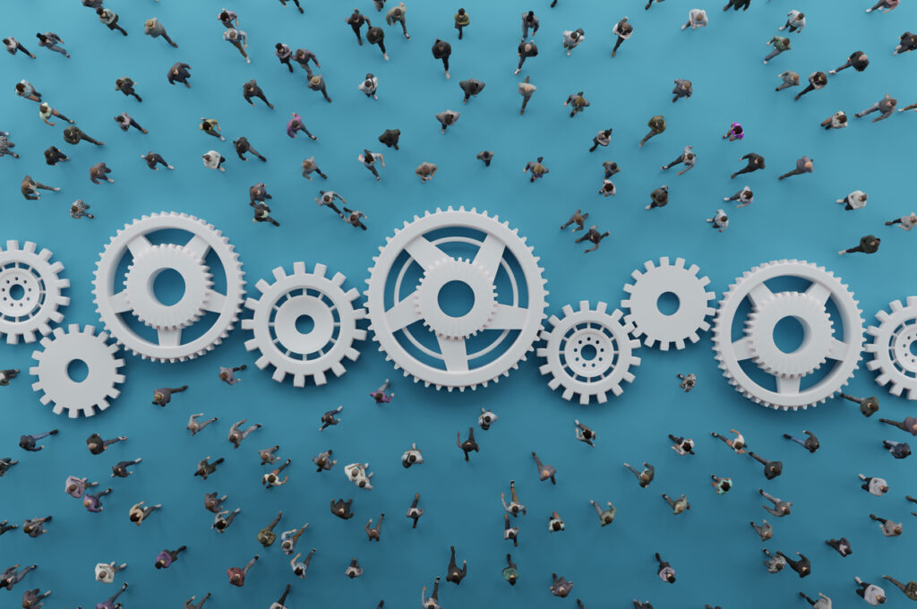 Overhead view of a diverse crowd walking across a bright blue surface interlinked with large white gears. The mechanical cogs symbolize teamwork, human systems, and the power of collective collaboration. Ideal for business, HR, and innovation themes.
