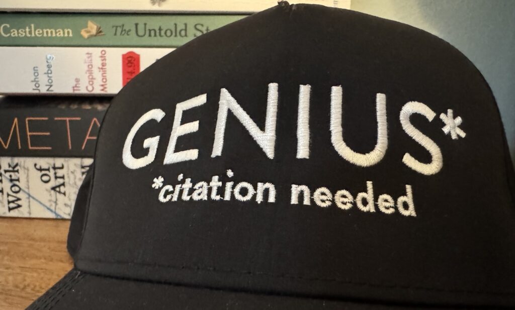 A black baseball cap with white lettering that reads "GENIUS* - Citation Needed" set on a table in front of a stack of books