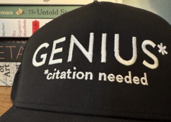 A black baseball cap with white lettering that reads "GENIUS* - Citation Needed" set on a table in front of a stack of books