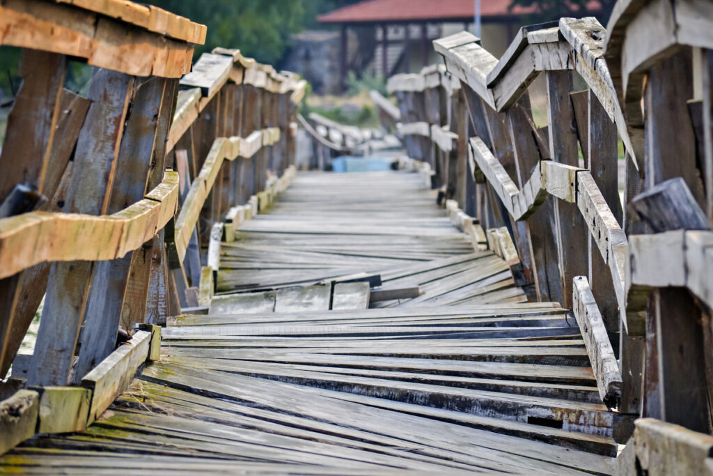 Decorative image representing a dangerous path ahead. Closeup of a deformed and dilapidated wooden bridge over the small river. Weathered and broken wood planks of a very old and damaged pedestrian bridge near the village.