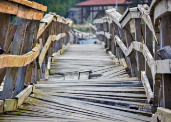Decorative image representing a dangerous path ahead. Closeup of a deformed and dilapidated wooden bridge over the small river. Weathered and broken wood planks of a very old and damaged pedestrian bridge near the village.