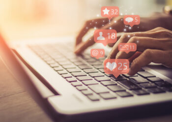 hands typing on a laptop keyboard with social media icons such as likes superimposed over the image