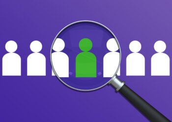 A decorative image of a magnifying glass highlights a unique green Icon from a crowd of figures, symbolizing the Recruitment process and Headhunting - Recruitment. This powerful visual represents Finding the right Employee and recognizing Individuality and potential. It's an ideal Concept for Human Resources, illustrating the Focus - Concept and Scrutiny required to build a strong team and identify future Leadership.